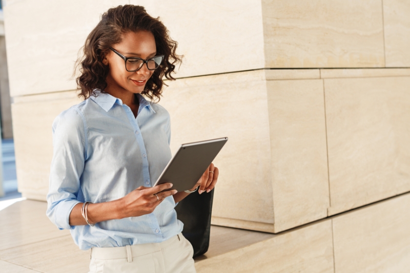 A woman wearing glasses and a light blue shirt stands outdoors reading a tablet, smiling slightly.